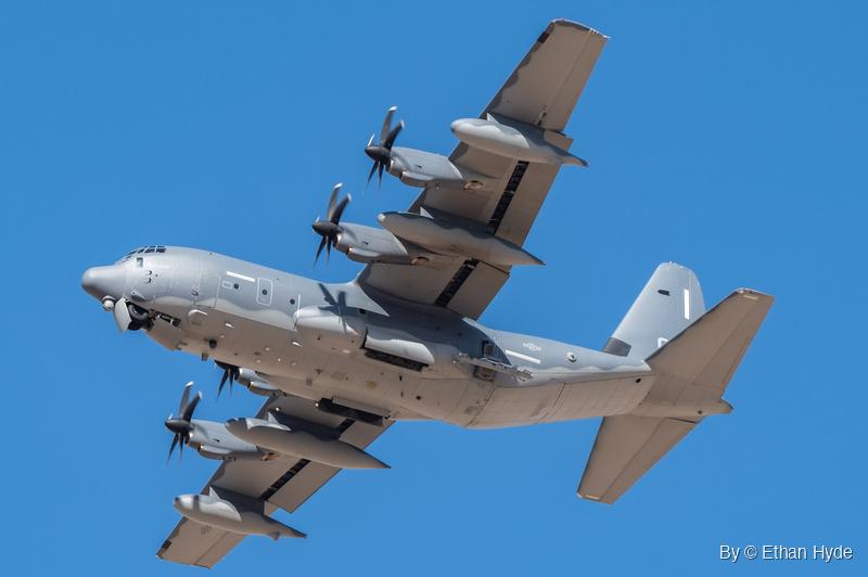 A gray four-engine military turboprop with six-bladed propellers, underwing fuel tanks and outer-wing pods passes overhead against clear blue sky.