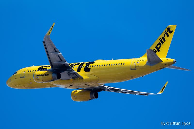 A yellow Spirit Airlines Airbus A320 rolls into a steep bank, seen from below with one wing raised against deep blue sky.