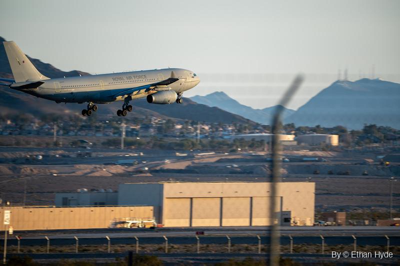 A Royal Air Force A330 Voyager tanker lands with gear down above a hangar and perimeter fence, with houses and desert mountains behind.