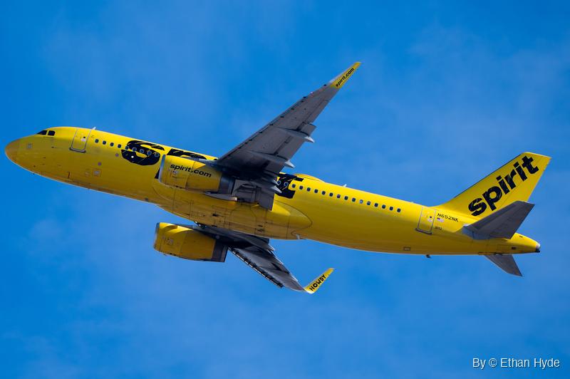 A bright yellow twin-engine airliner climbs away nose-up with its gear retracted, seen from below against blue sky and thin cloud.