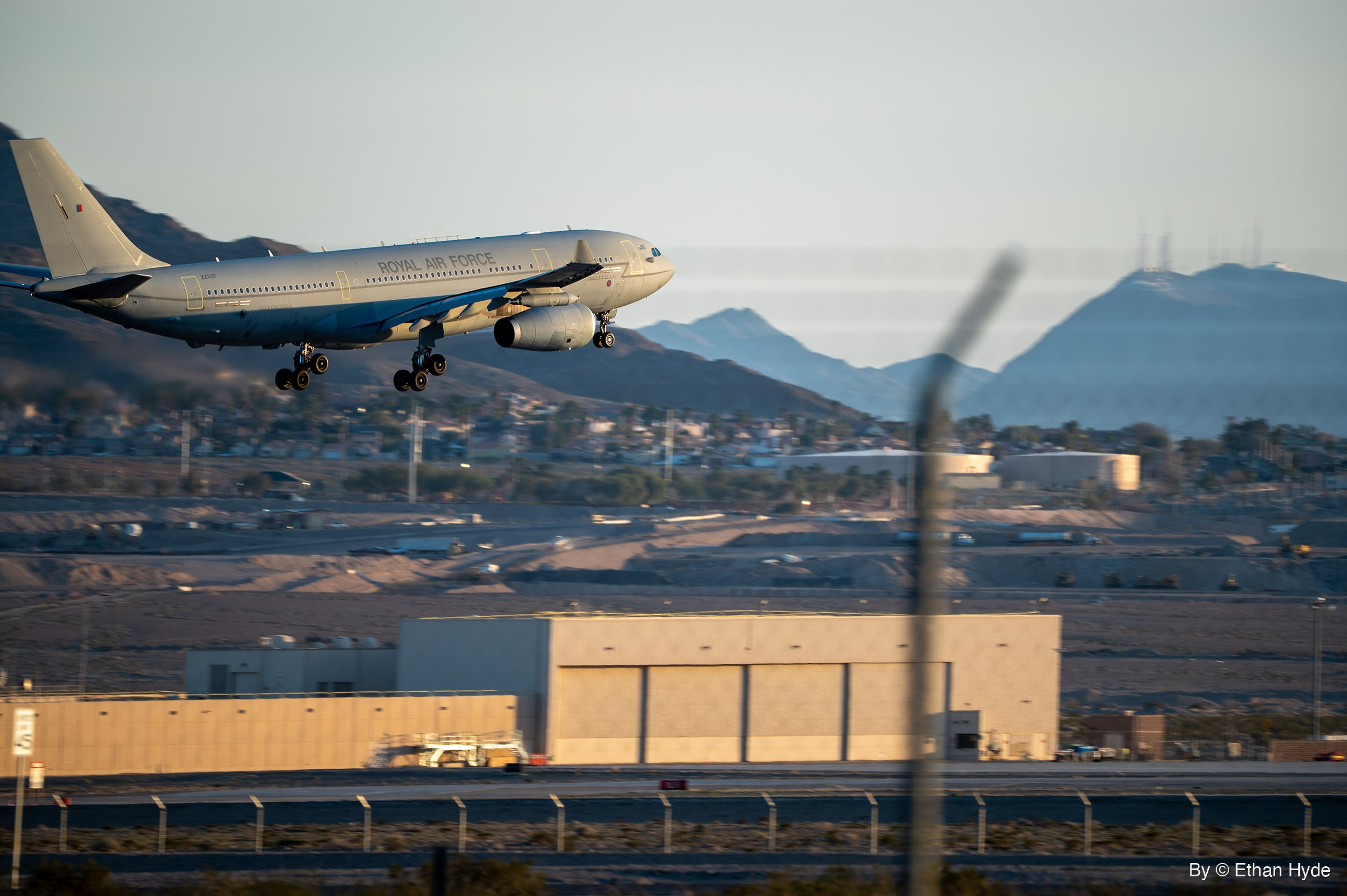 A Royal Air Force A330 Voyager tanker lands with gear down above a hangar and perimeter fence, with houses and desert mountains behind.