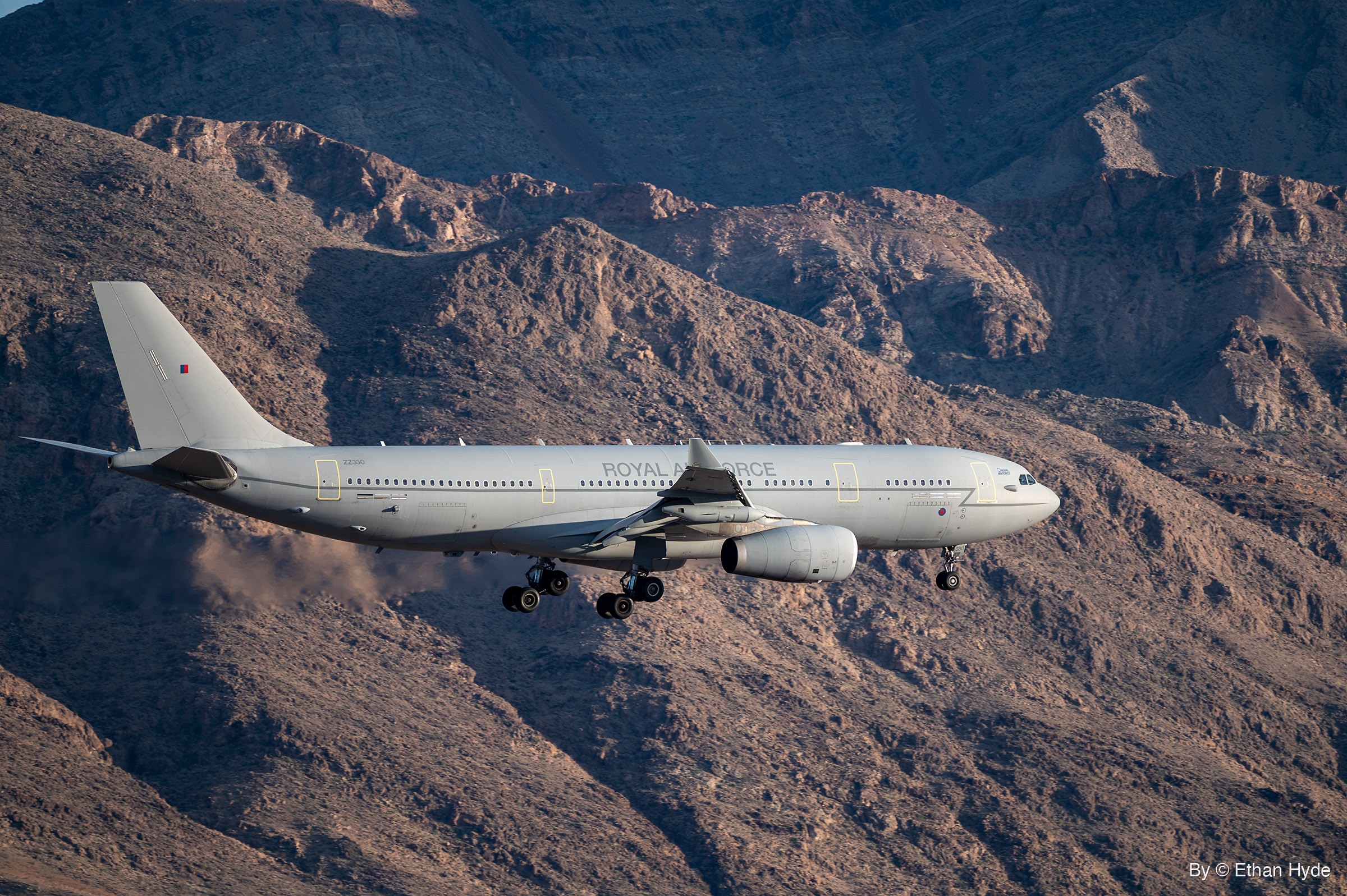 A gray Royal Air Force Voyager tanker flies gear-down past rugged brown desert mountains on approach.