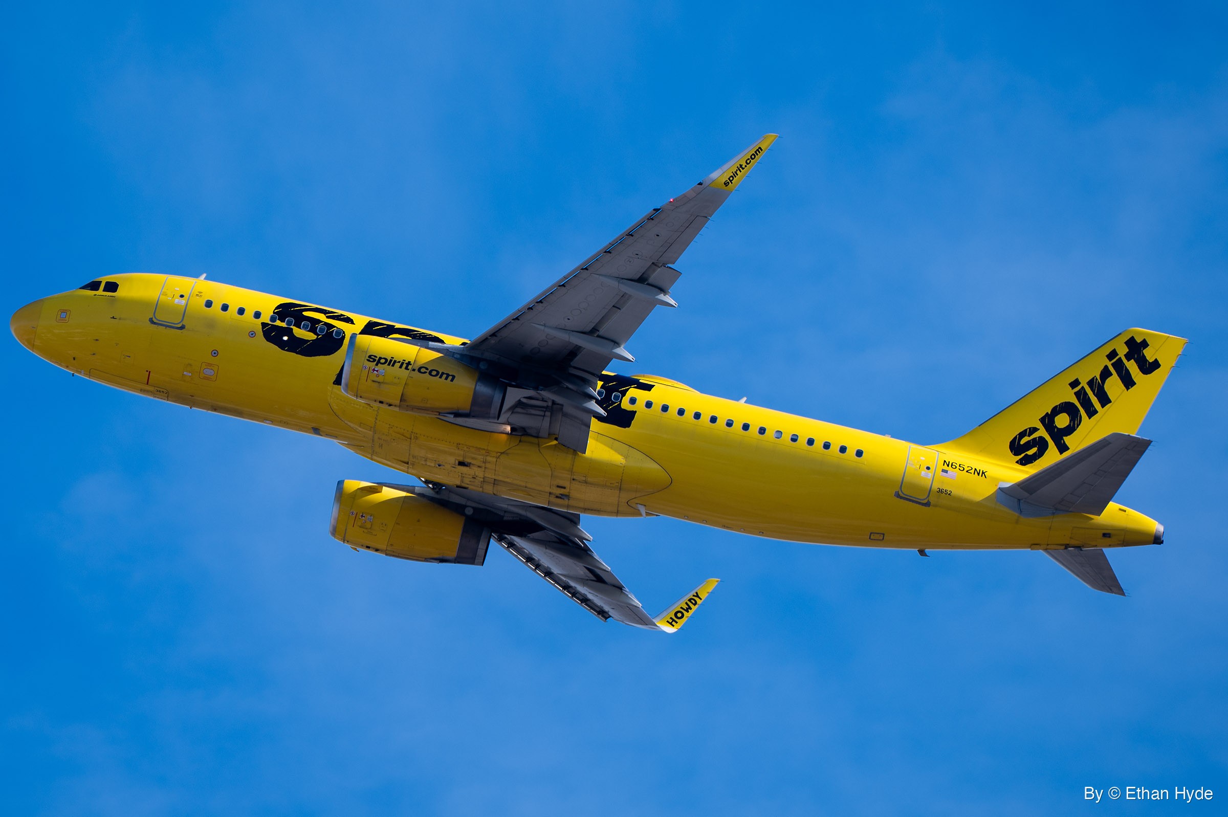 A bright yellow twin-engine airliner climbs away nose-up with its gear retracted, seen from below against blue sky and thin cloud.