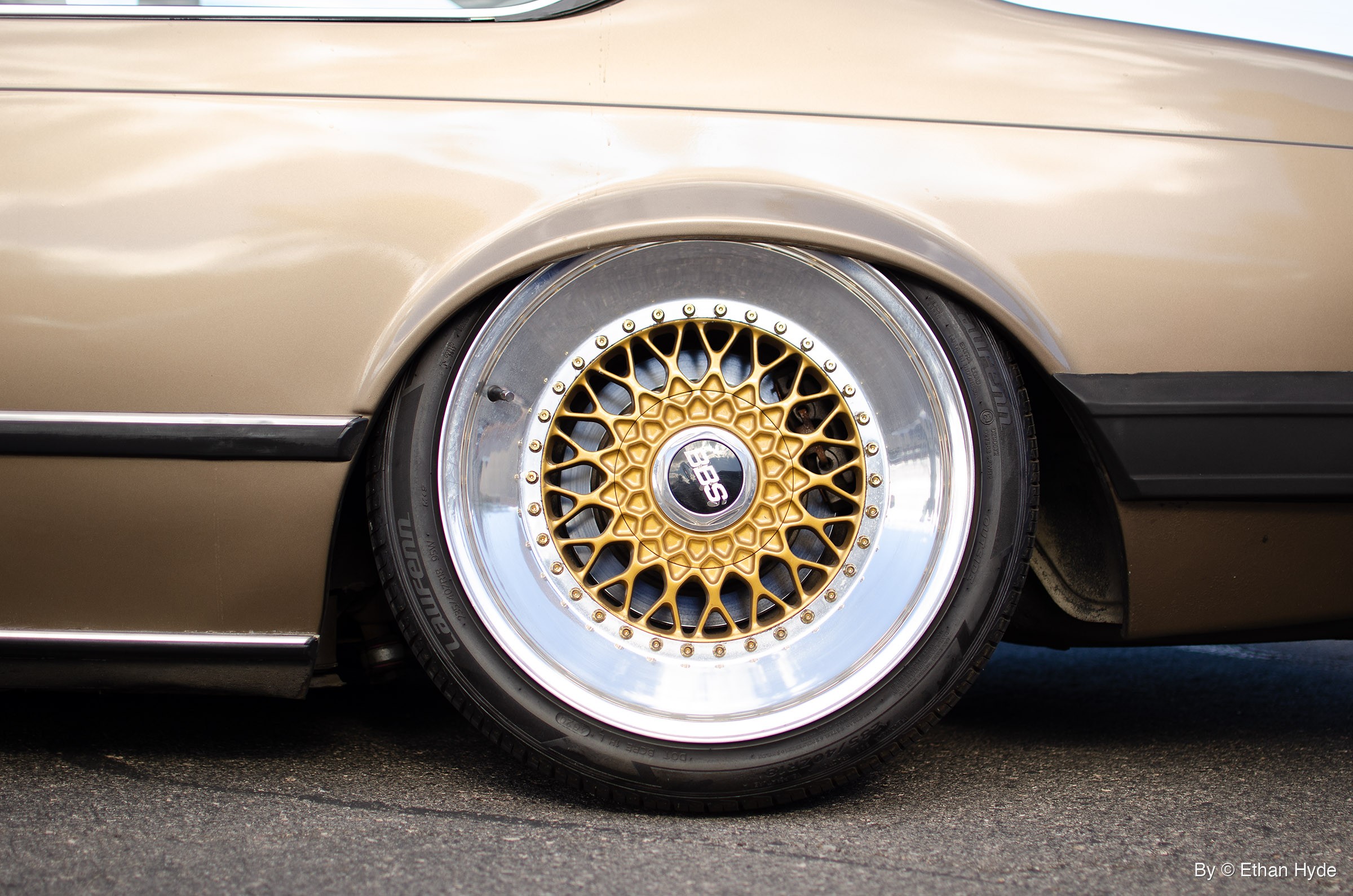 Close-up of a gold mesh wheel with a wide polished lip and a ring of small rim bolts under the rear fender of a bronze coupe.