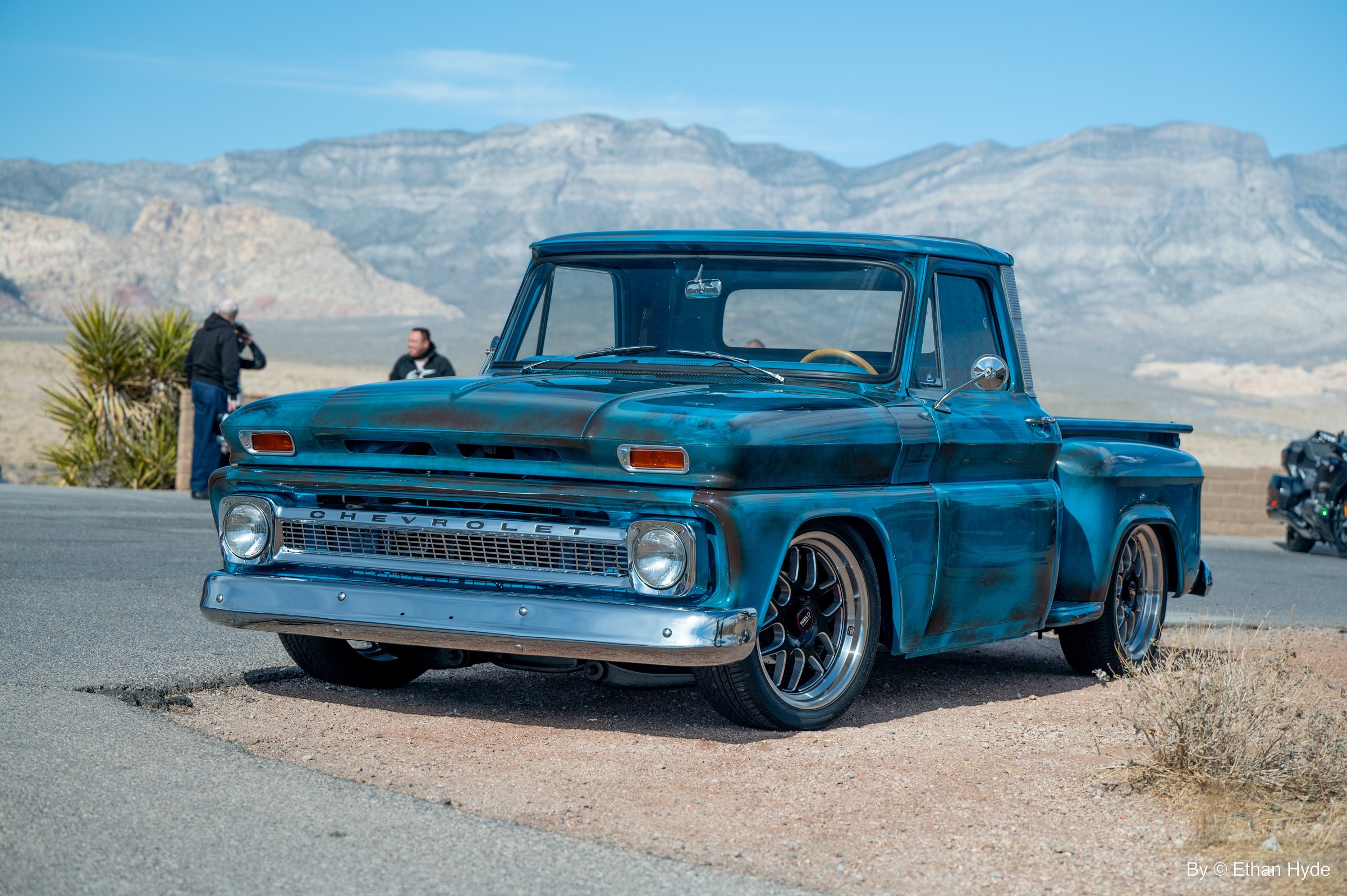 A turquoise 1960s Chevrolet pickup on a desert road below red rock cliffs.