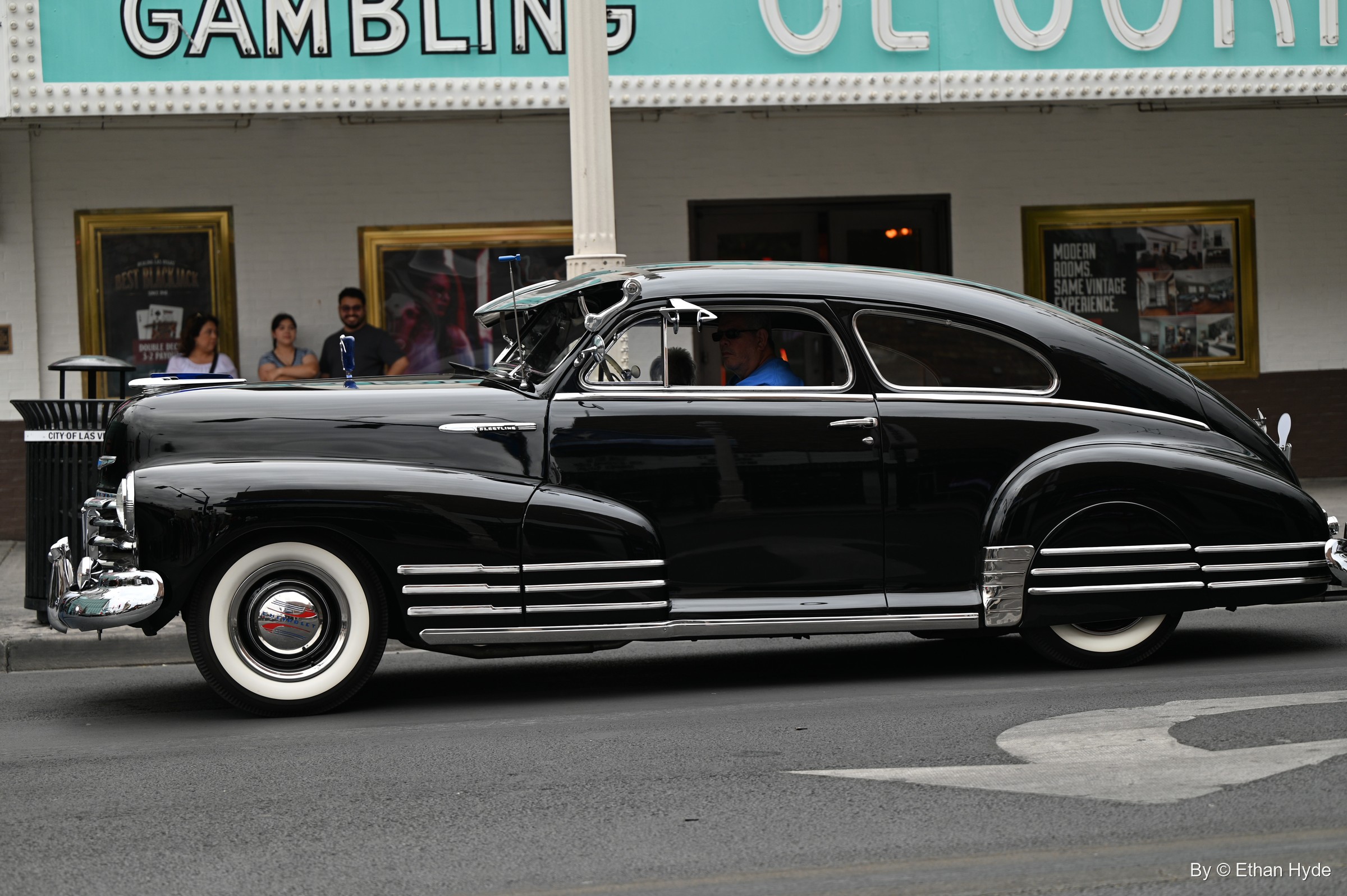 A black 1940s fastback sedan with chrome trim and whitewall tires rolls past a casino frontage with a teal gambling sign.