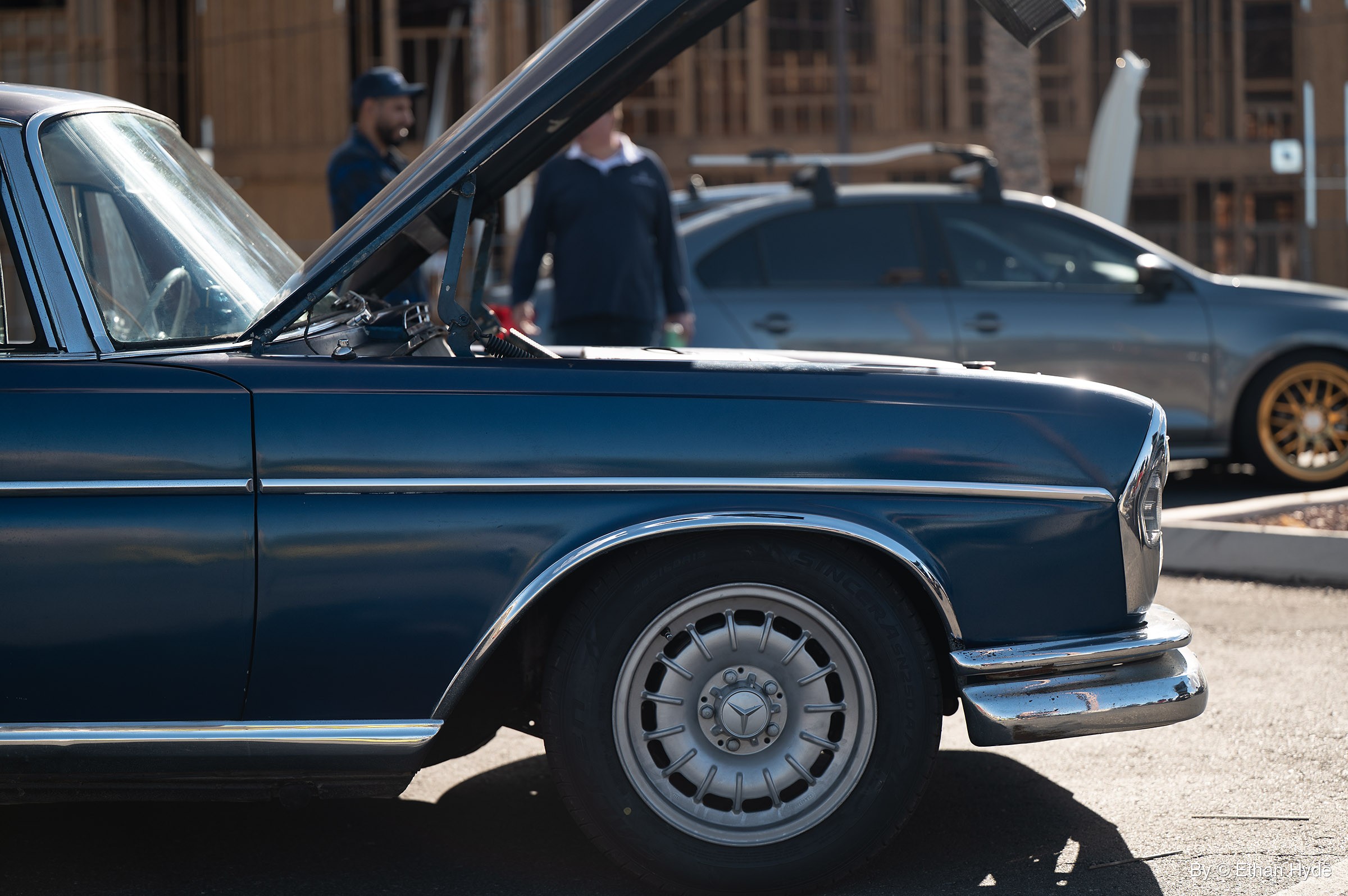 Front half of a dark blue vintage Mercedes 190 sedan with its hood raised at a car meet, two men and a gray sedan behind.