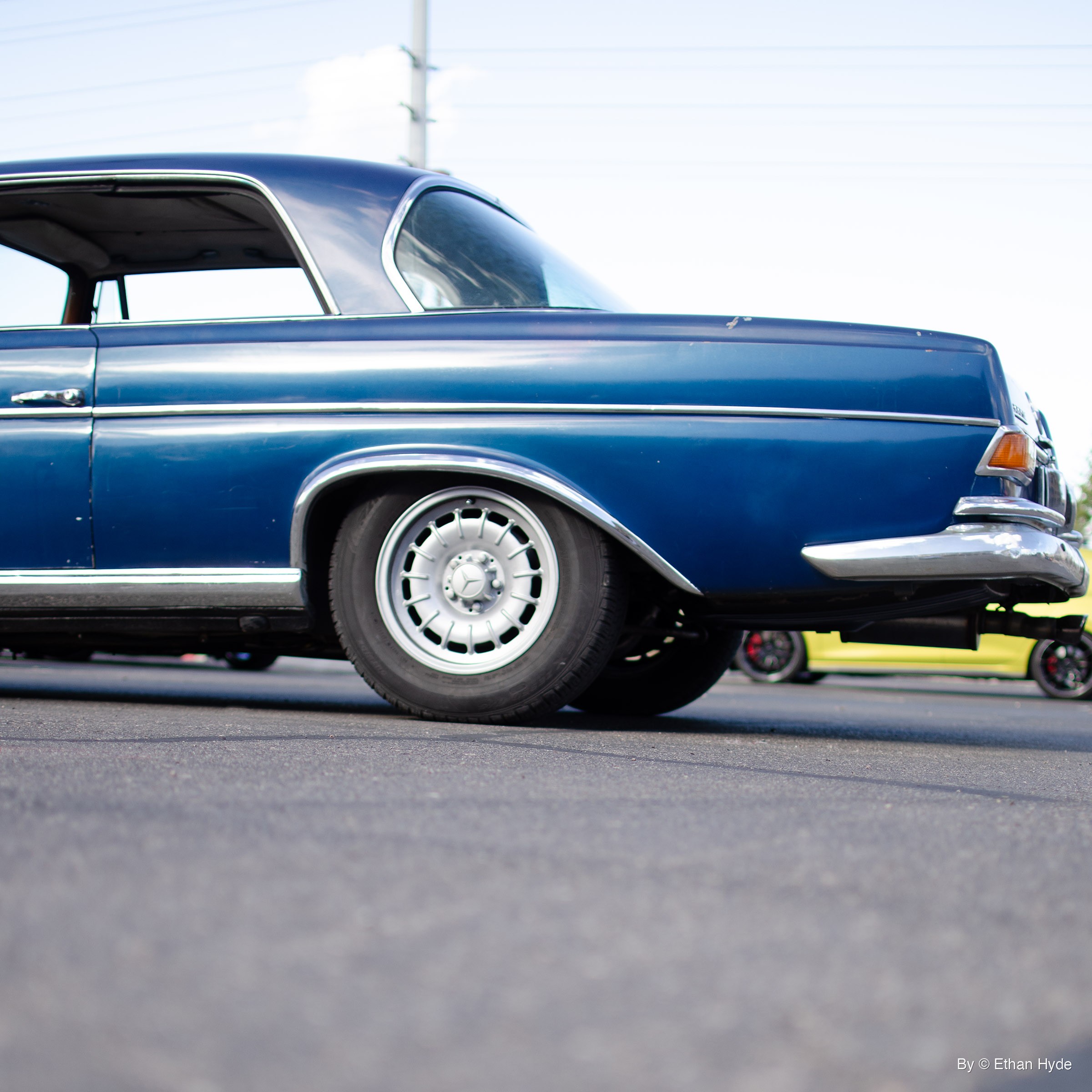 Low ground-level view of the rear half of a dark blue Mercedes-Benz 280SE coupe with chrome trim and steel wheels, parked on asphalt.