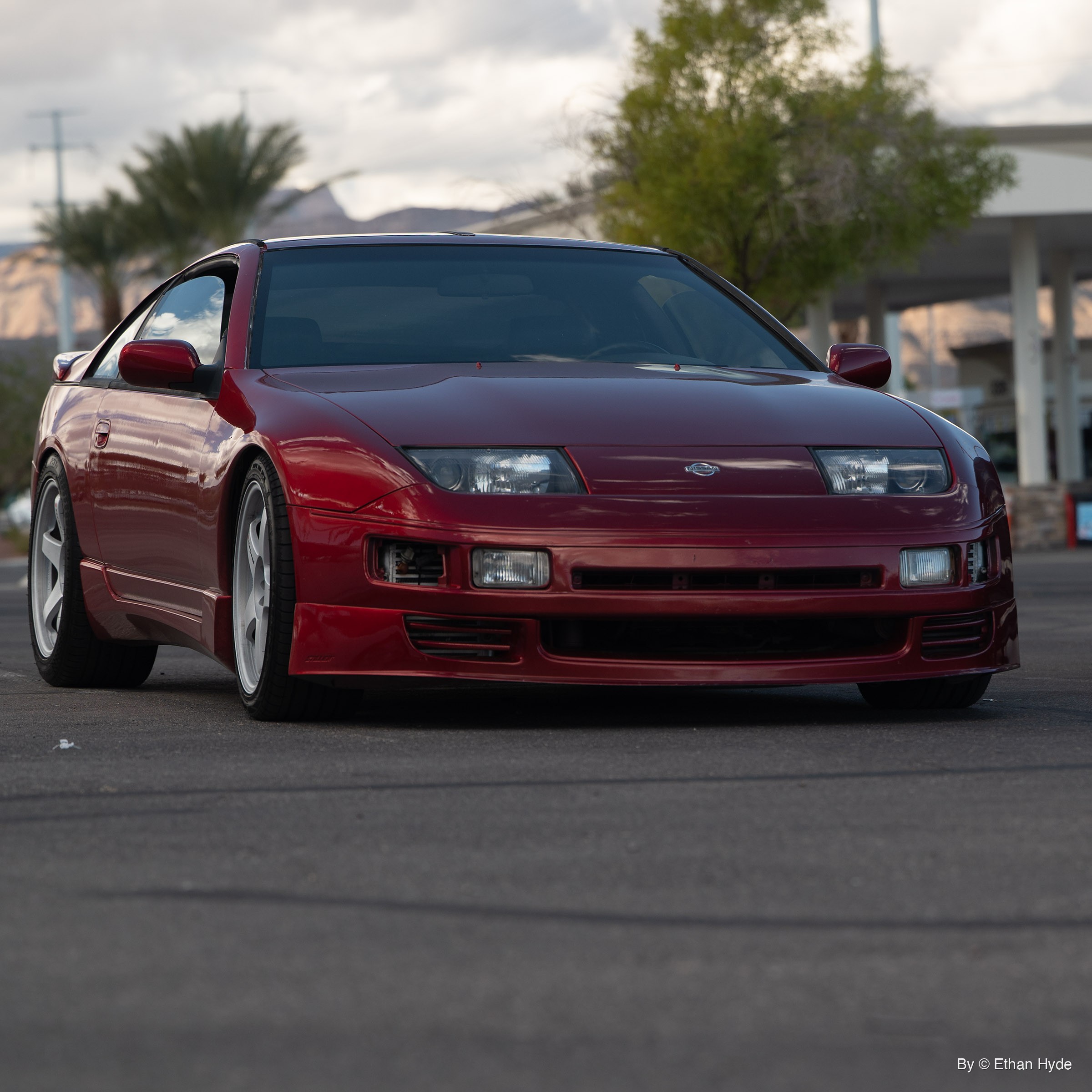 Deep red Nissan 300ZX with white wheels parked on asphalt, shot low from the front corner, palm trees and desert mountains behind.