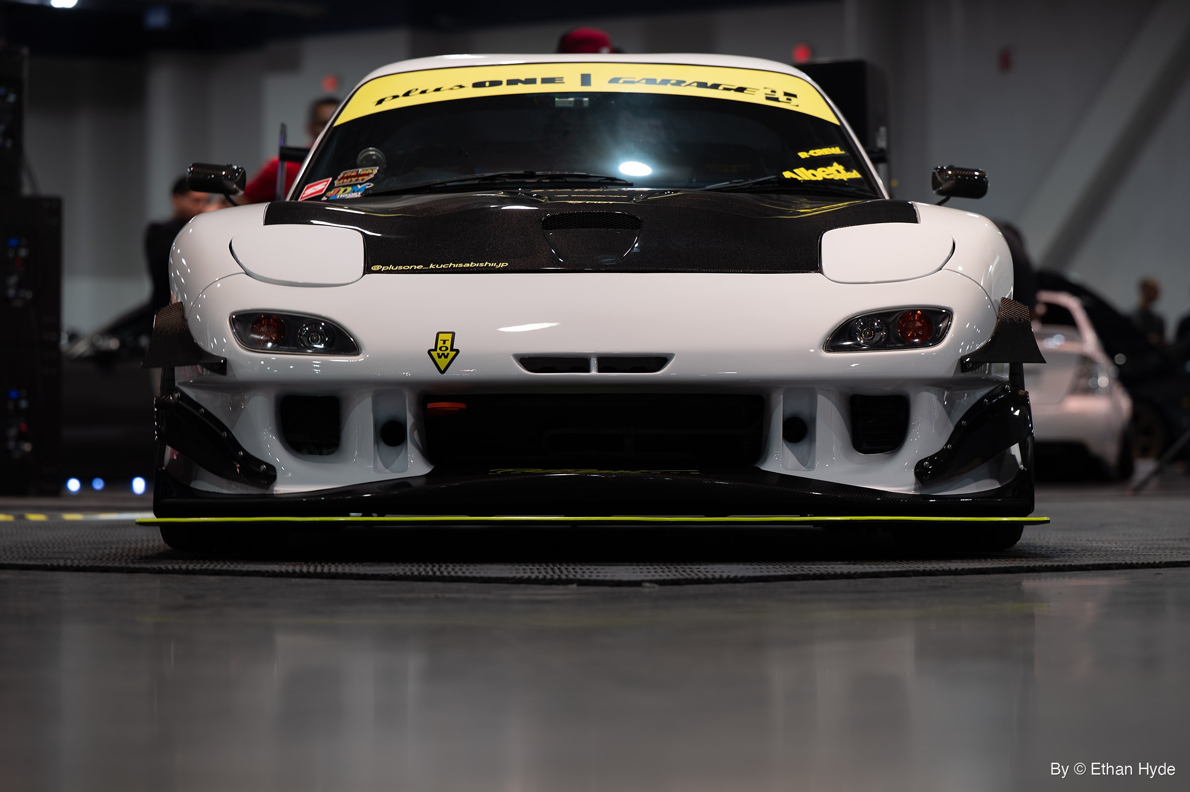 White Mazda RX-7 shot head-on at bumper height on an indoor show floor, with a carbon hood and a lime-green splitter edge.