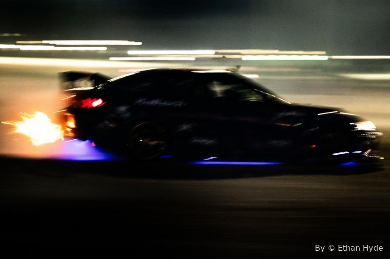 A car at night throwing flame from its exhaust, underglow on the pavement, mid-motion.