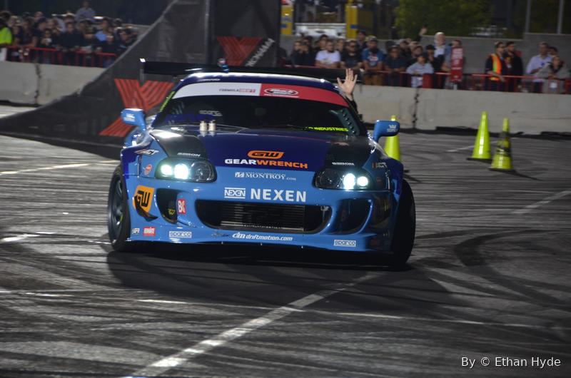 A blue liveried Toyota Supra drifting on a track, smoke off the rear tires.