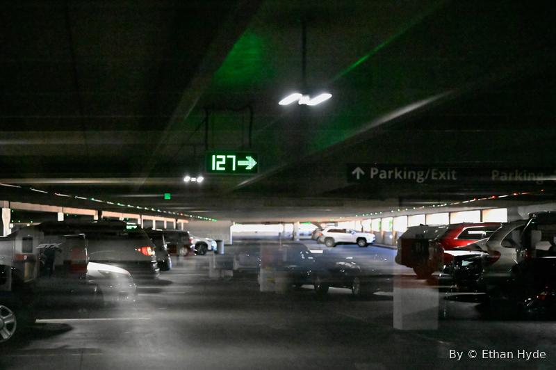 A dim parking garage at night lit by a green laser, cars and a level sign in the haze.
