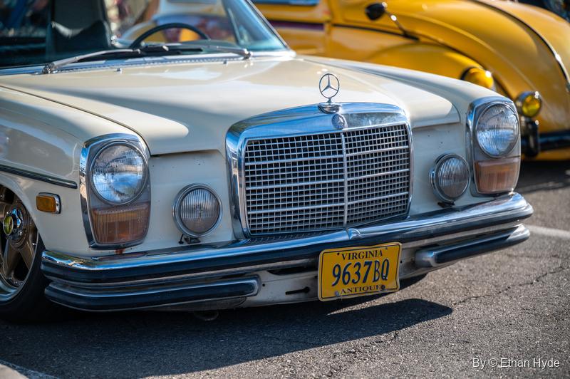 Front of a cream 1970s Mercedes-Benz sedan with a chrome grille and round headlights, a yellow car parked beside it.
