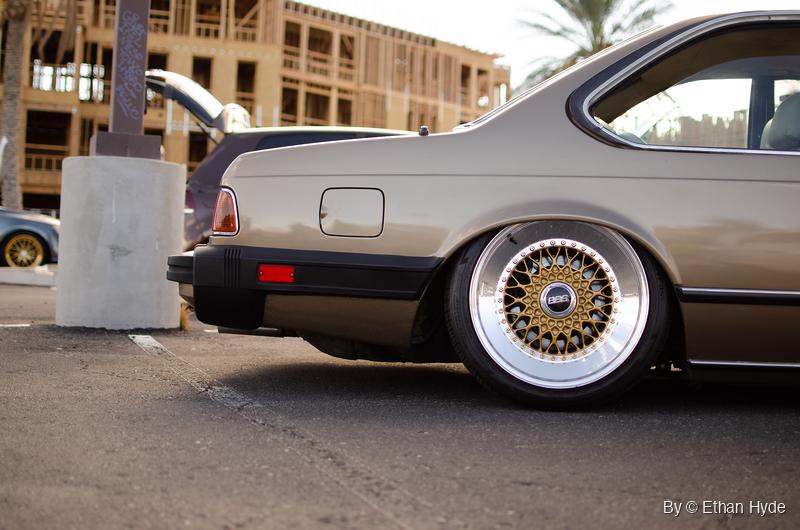 Rear three-quarter view of a lowered bronze BMW E24 coupe on gold mesh wheels with polished lips, parked beside a concrete post base.
