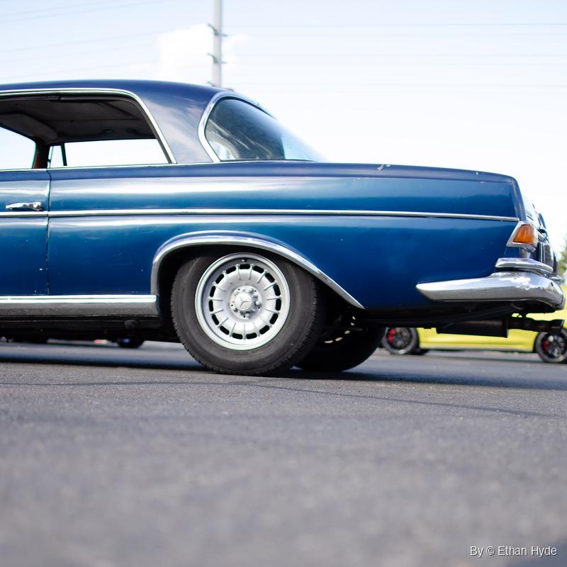 Low ground-level view of the rear half of a dark blue Mercedes-Benz 280SE coupe with chrome trim and steel wheels, parked on asphalt.