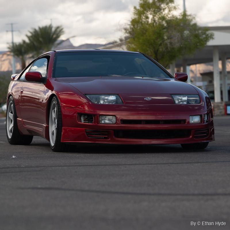 Deep red Nissan 300ZX with white wheels parked on asphalt, shot low from the front corner, palm trees and desert mountains behind.