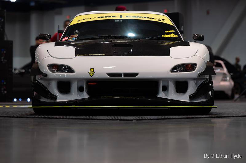 White Mazda RX-7 shot head-on at bumper height on an indoor show floor, with a carbon hood and a lime-green splitter edge.