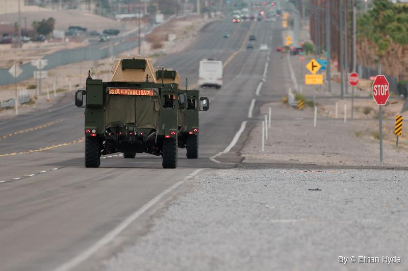 Two green armored vehicles with tan roof turrets drive away down a wide desert road, a student driver sign across the back of the nearer one.