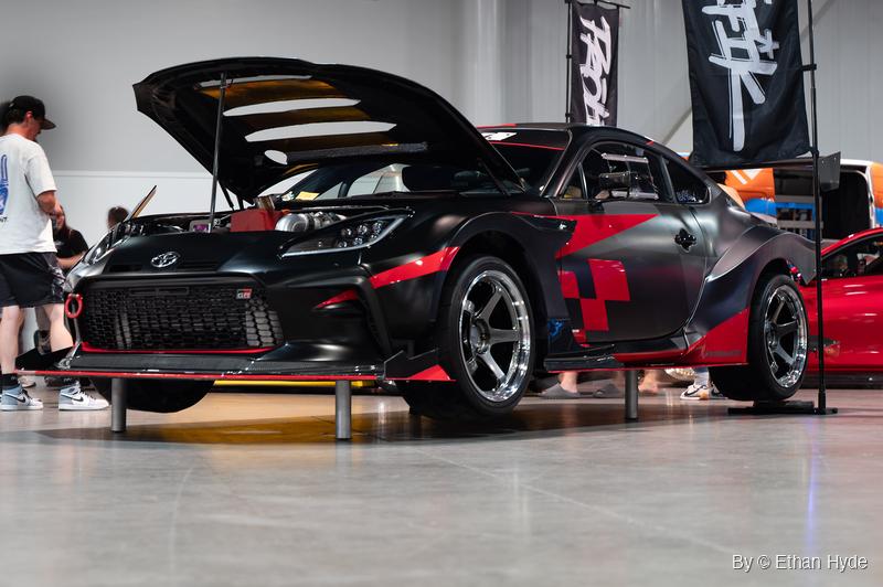 A black and red widebody Toyota GR86 with its vented hood propped open stands on metal posts at an indoor car meet as a visitor looks on.
