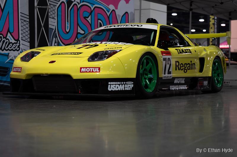 A yellow, heavily modified Honda NSX race car with green wheels sits low on a polished concrete floor at an indoor car show.