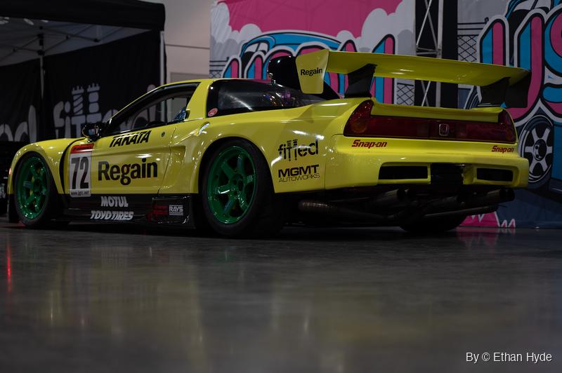 Rear three-quarter view of a yellow Honda NSX race car with a tall rear wing and green wheels, parked before a pink and blue mural backdrop.