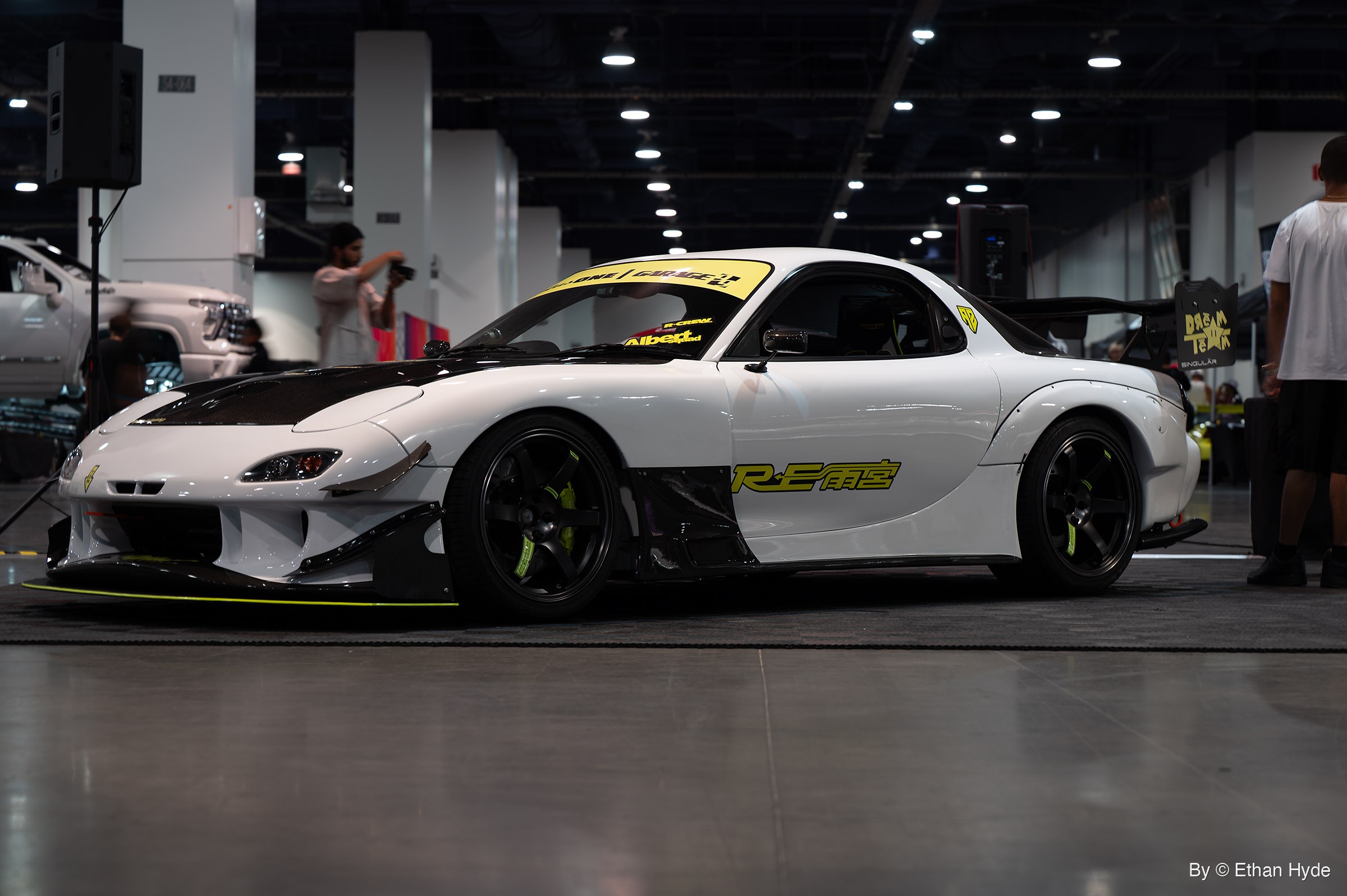 White widebody Mazda RX-7 with a black hood, black wheels and a rear wing, seen from the side on an indoor show floor.