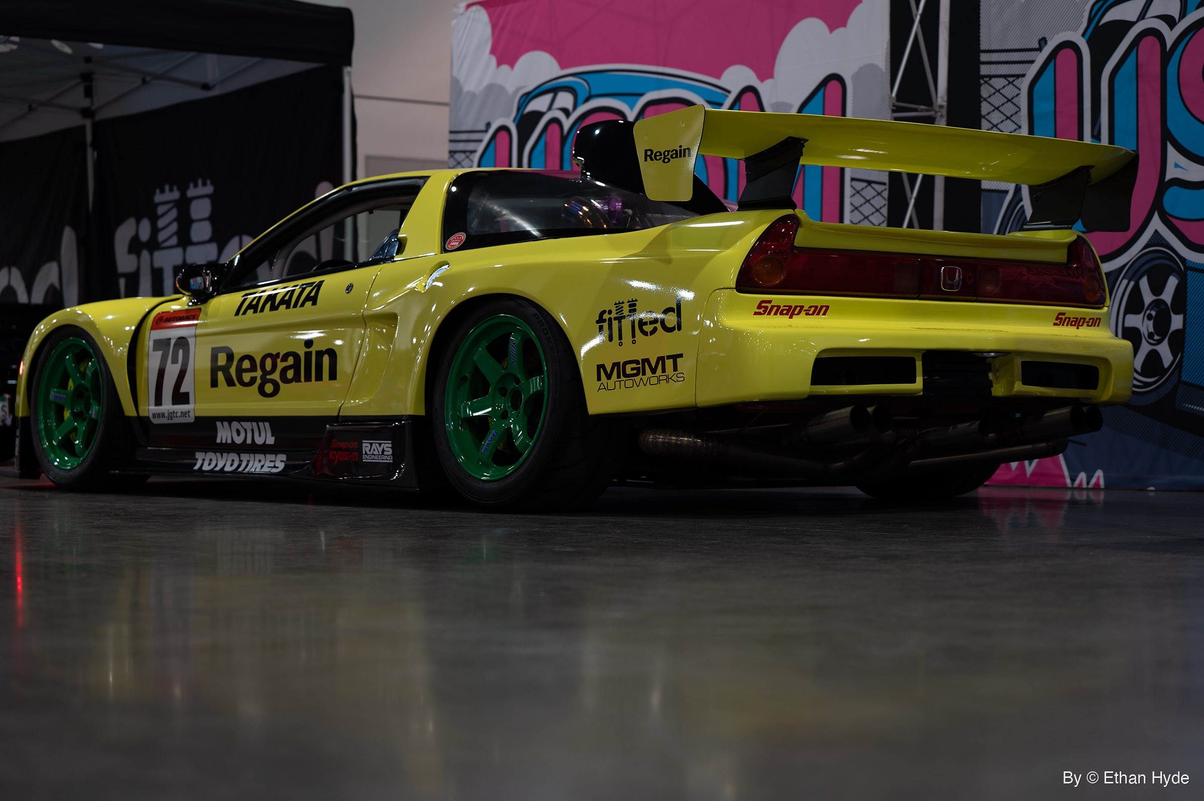 Rear three-quarter view of a yellow Honda NSX race car with a tall rear wing and green wheels, parked before a pink and blue mural backdrop.