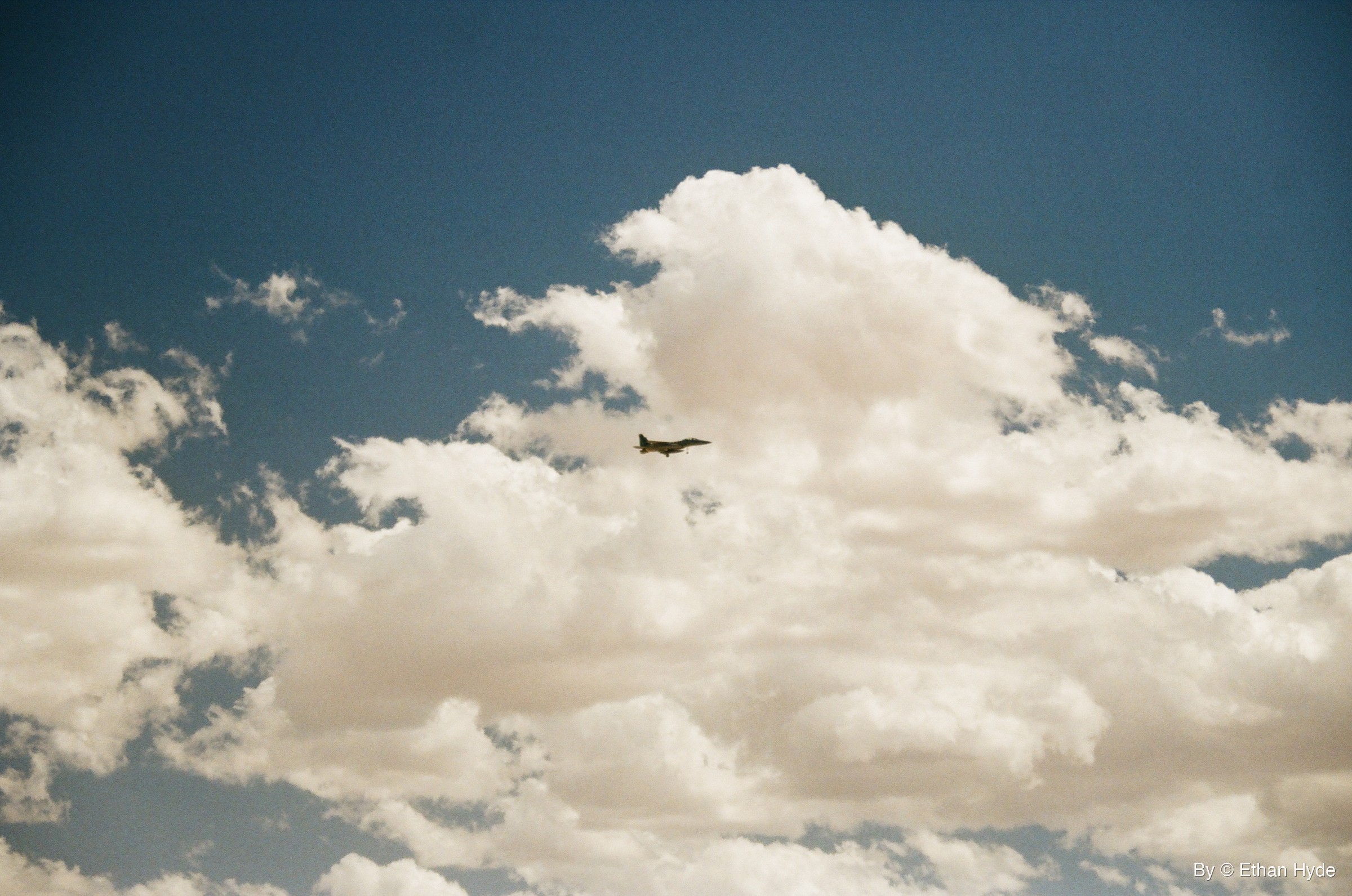 A fighter jet above a bank of white cumulus clouds, shot on film.