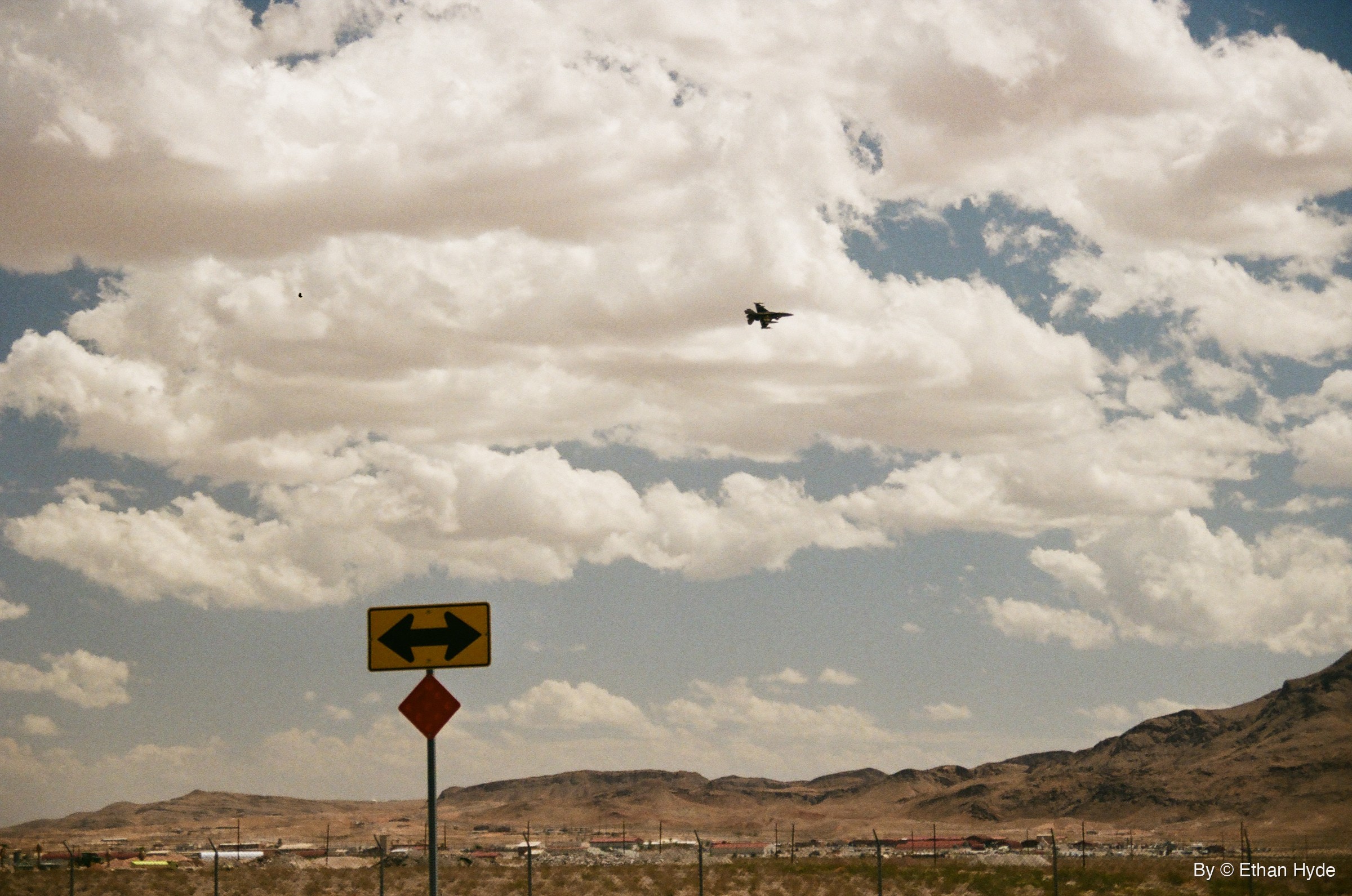 A fighter jet flying above a brown desert ridge under scattered clouds, shot on film.