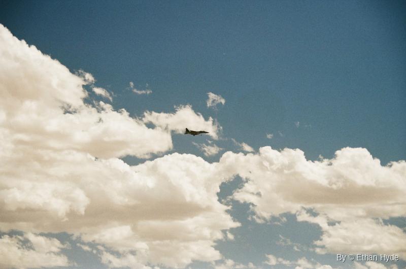 A fighter jet passing between bright cumulus clouds in a deep blue sky, shot on film.