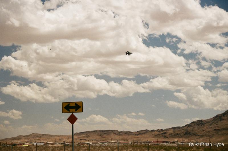 A fighter jet flying above a brown desert ridge under scattered clouds, shot on film.