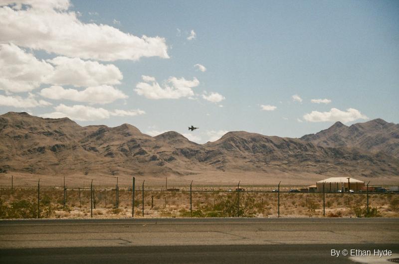 A fighter jet low over desert mountains beyond a fence line, shot on film.