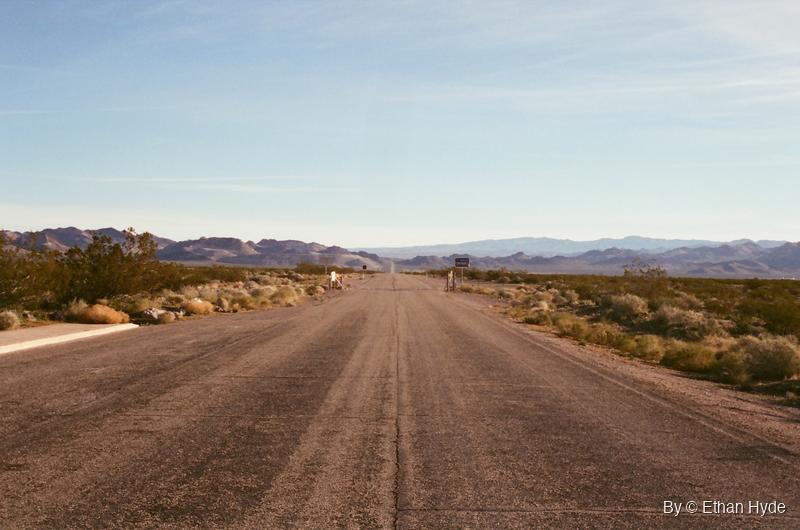 A straight dirt road crossing open desert toward low hills, shot on film.