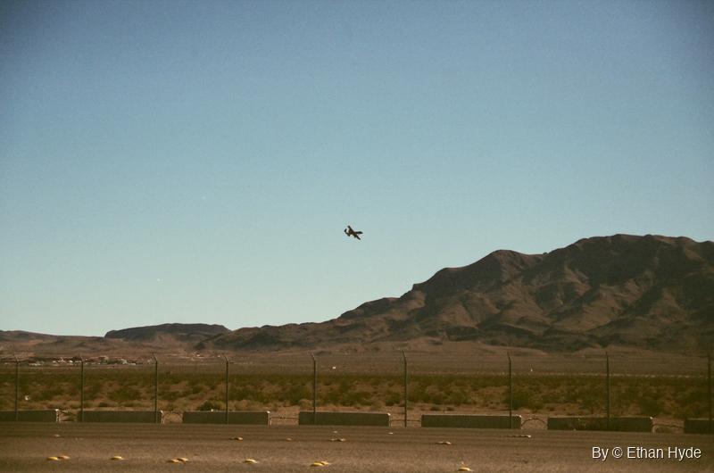 A distant jet above desert mountains seen beyond a wire fence, on film.