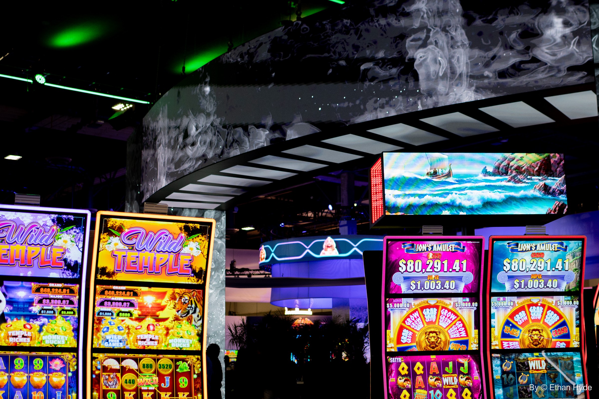 Slot machine cabinets in bright colors beneath a wide ocean-wave screen at a G2E booth.