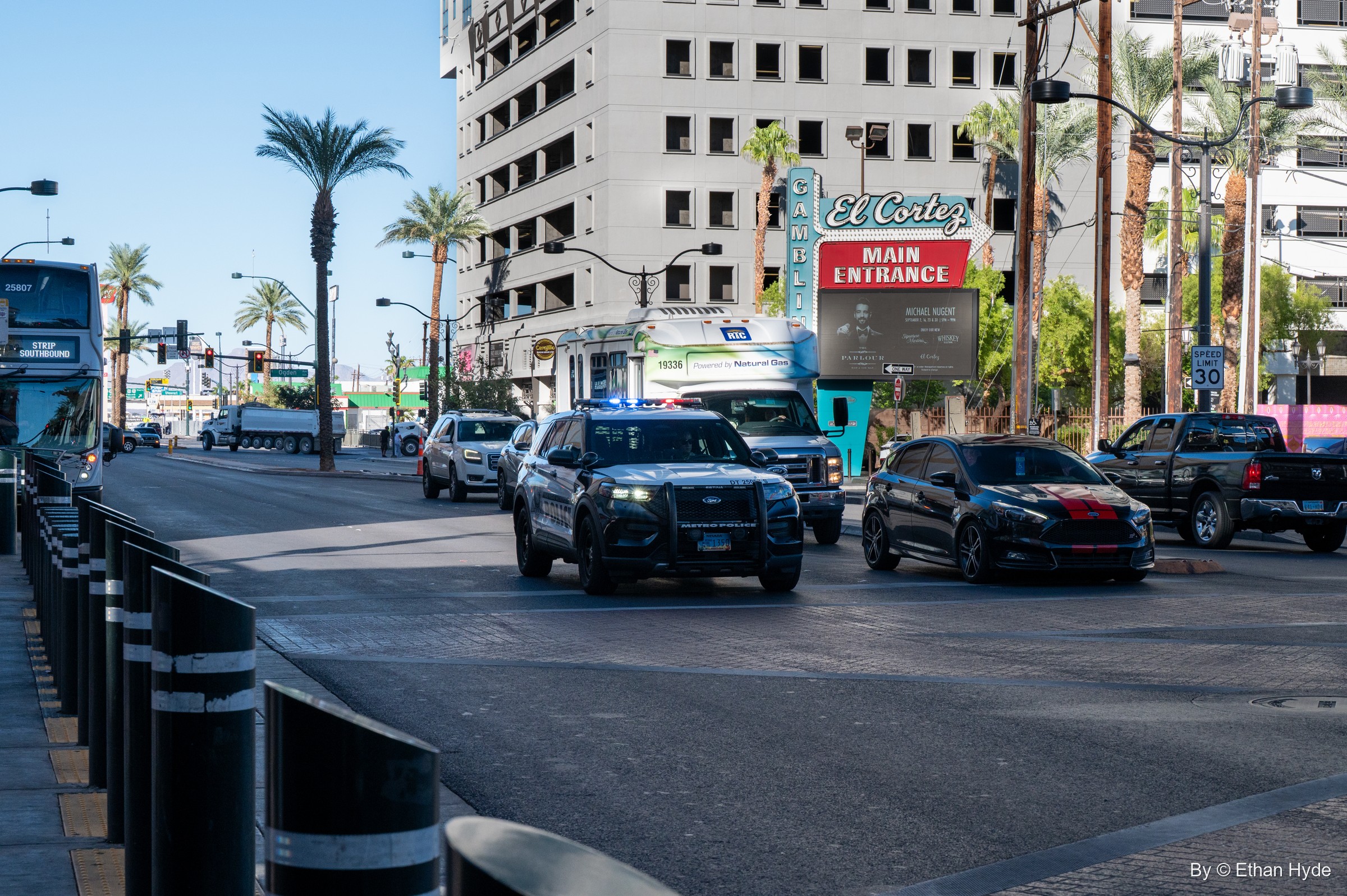 A police SUV and cars drive down a sunny downtown boulevard lined with palms, bollards and a casino marquee.