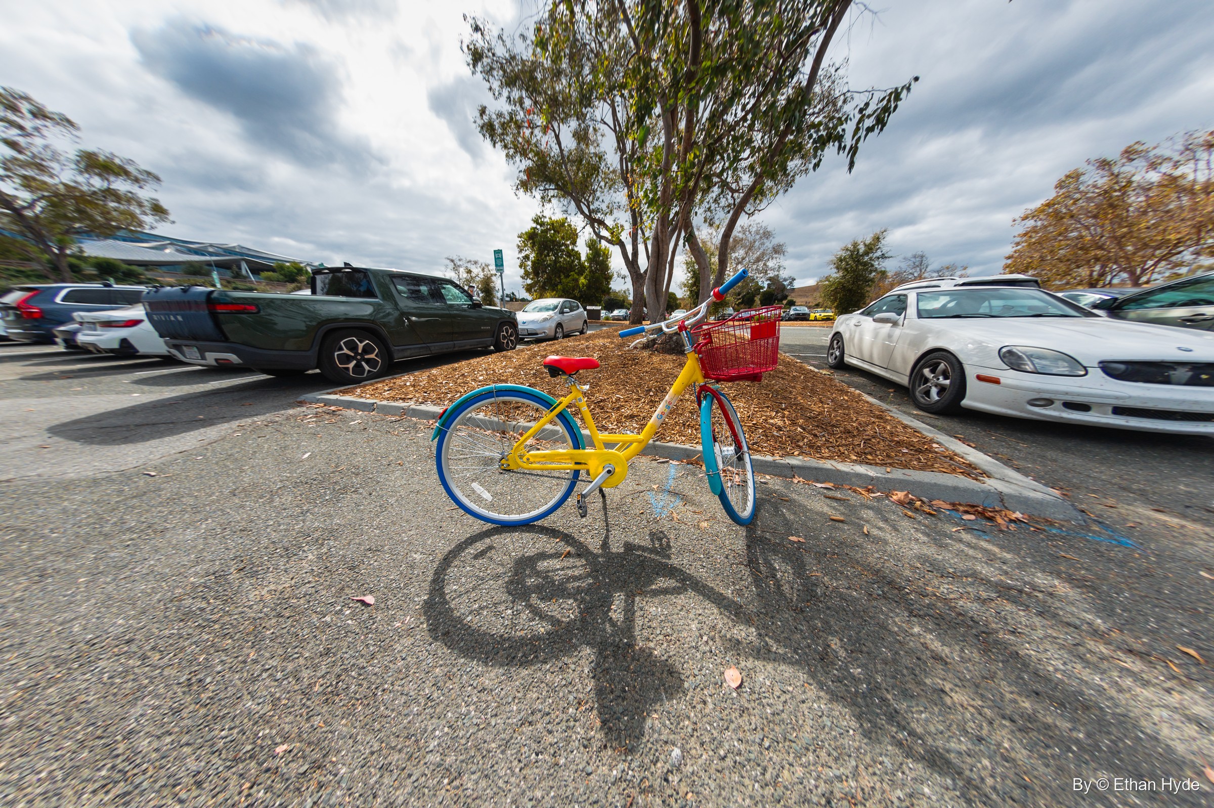 Close fisheye view of a yellow Google bike with a red basket casting a long shadow across a parking lot.