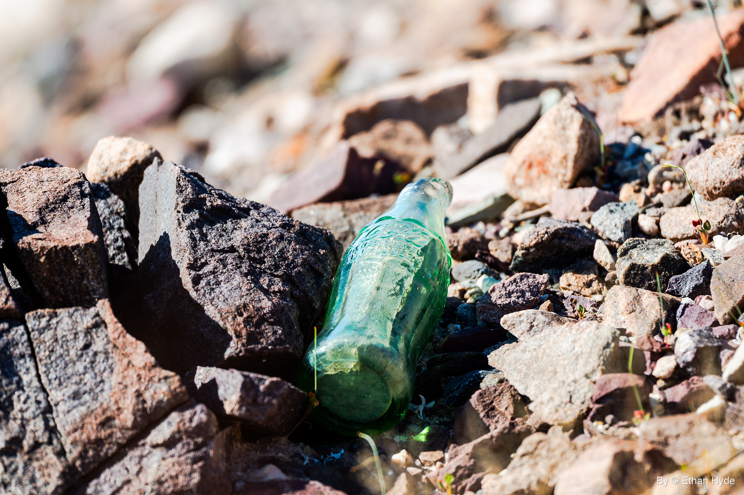 An old green glass soda bottle lies on its side, wedged between dark rocks and loose gravel in bright sun.