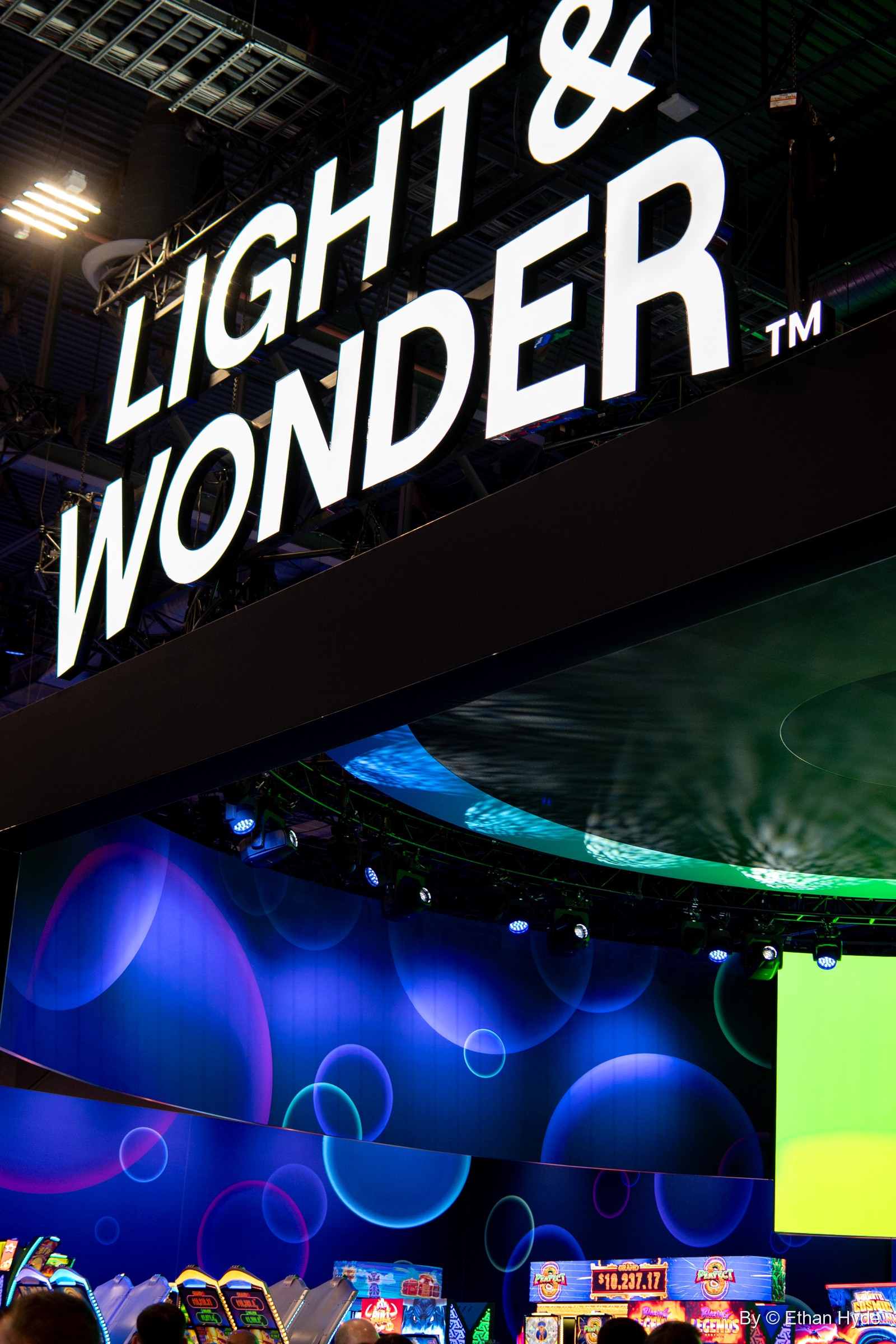 A white Light & Wonder sign above a booth of glowing blue and green screens on the G2E show floor.