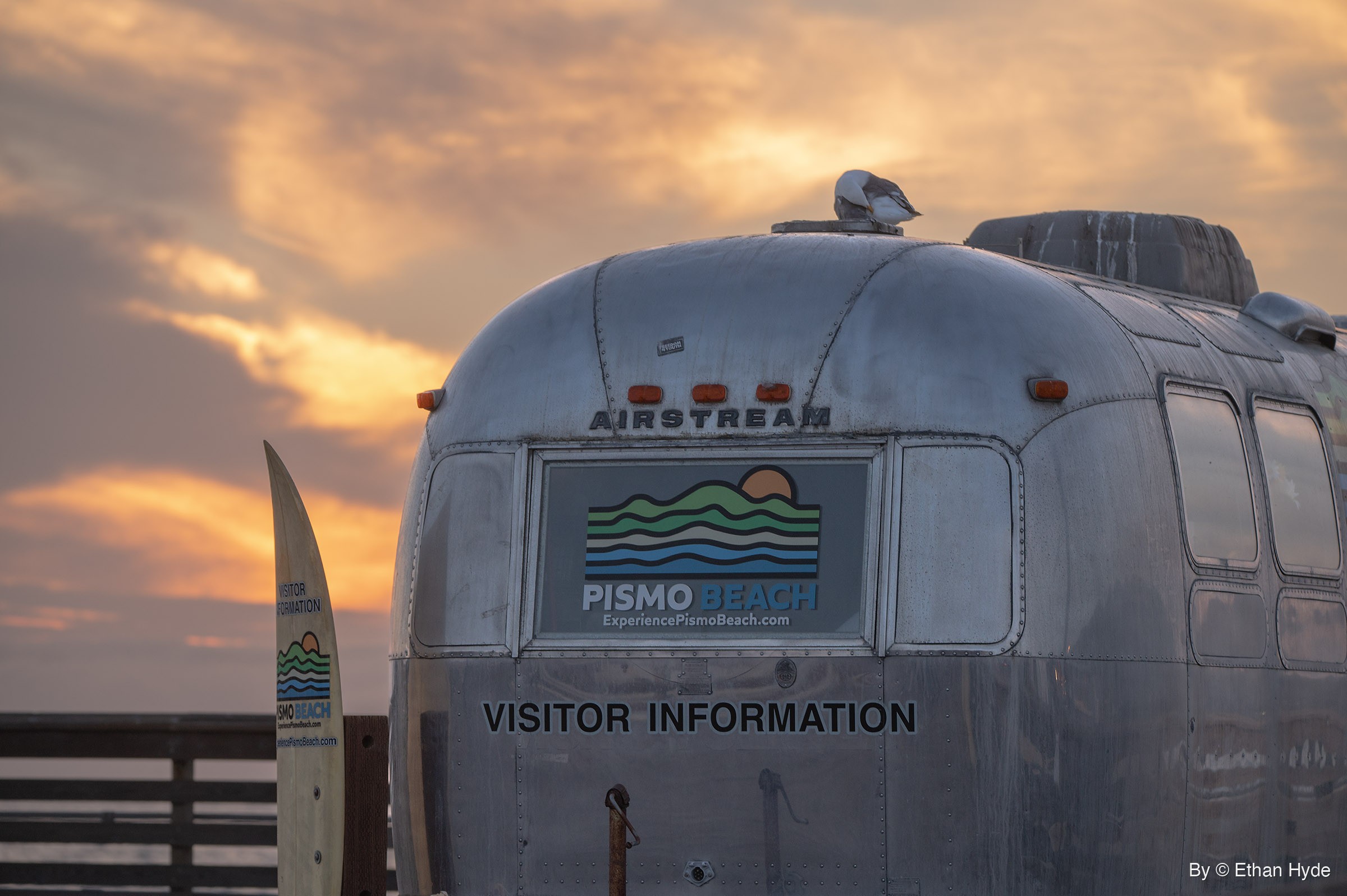 A polished aluminum Airstream trailer marked Visitor Information with a Pismo Beach logo, a gull on its roof and a surfboard sign beside it, under an orange sunset sky.