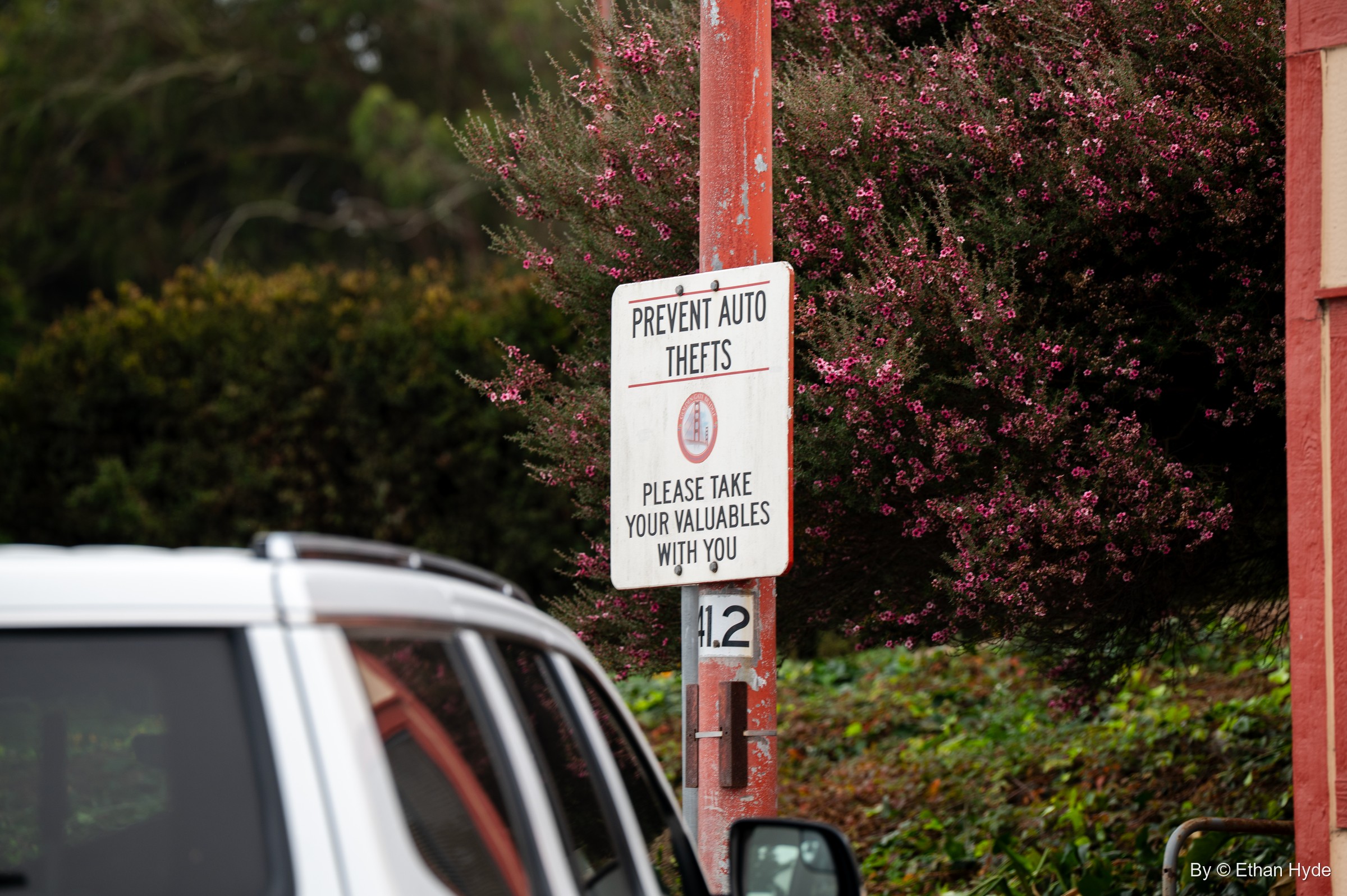 A red-and-white sign reading "Prevent auto thefts, please take your valuables with you" on a red post beside a parked SUV and flowering shrubs.