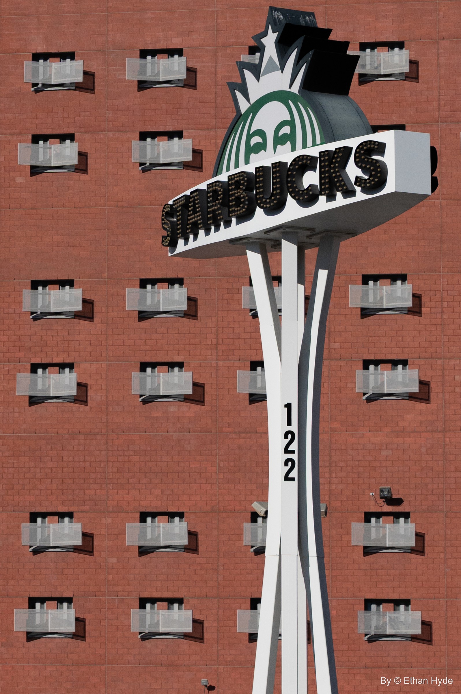 A Starbucks sign shaped like an observation tower on a white pole in front of a red brick wall of small windows.