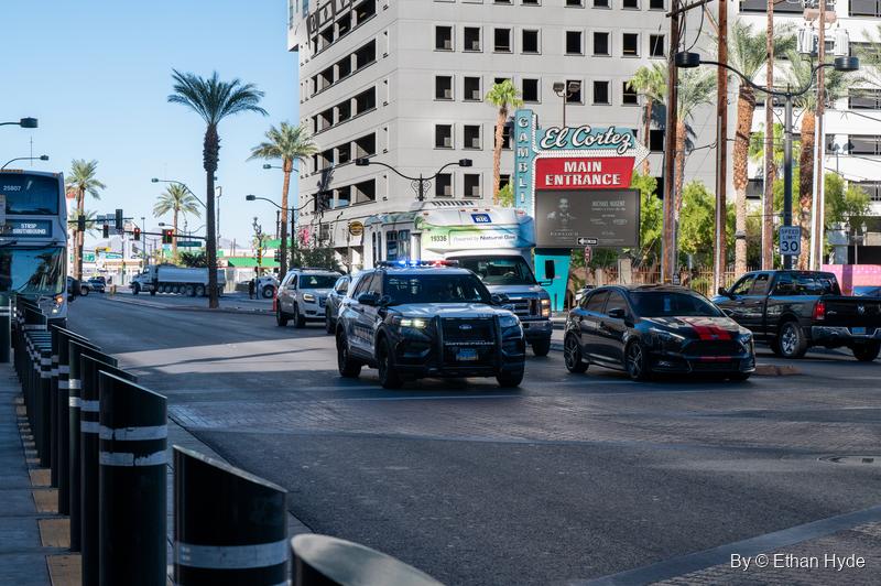 A police SUV and cars drive down a sunny downtown boulevard lined with palms, bollards and a casino marquee.