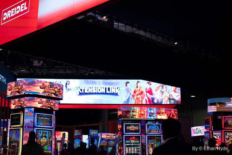 Rows of lit gaming cabinets under a long curved Extension Link screen and a Dreidel sign on the G2E floor.