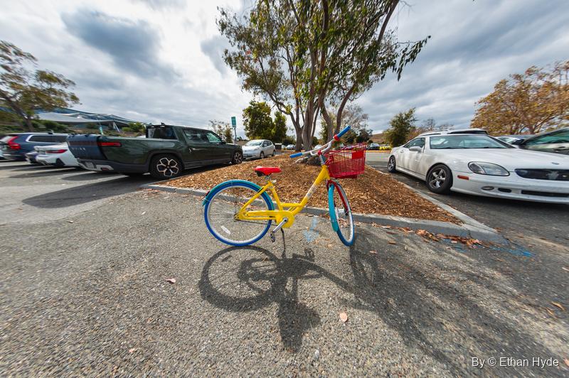 Close fisheye view of a yellow Google bike with a red basket casting a long shadow across a parking lot.