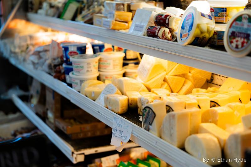 Stacked wheels and blocks of cheese on the shelves of a refrigerated case.