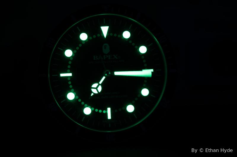 A dive watch face glowing green in the dark, hands and markers lit.