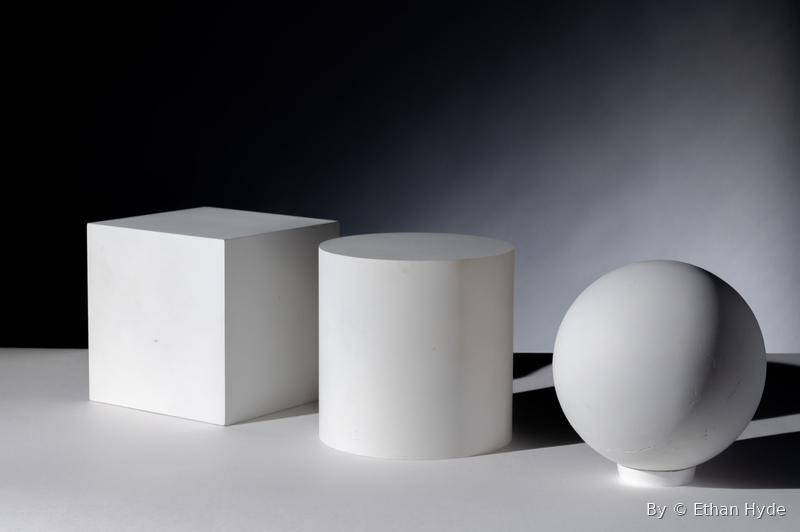 A studio still life of white geometric shapes — cube, cylinder, and sphere — under soft light.