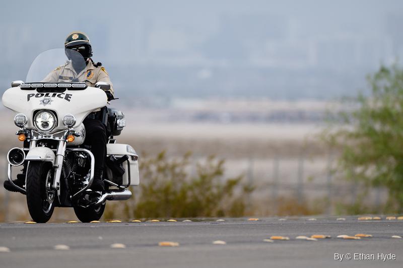 A uniformed motorcycle officer in a helmet and sunglasses rides a white touring motorcycle along a road lined with raised reflective markers.